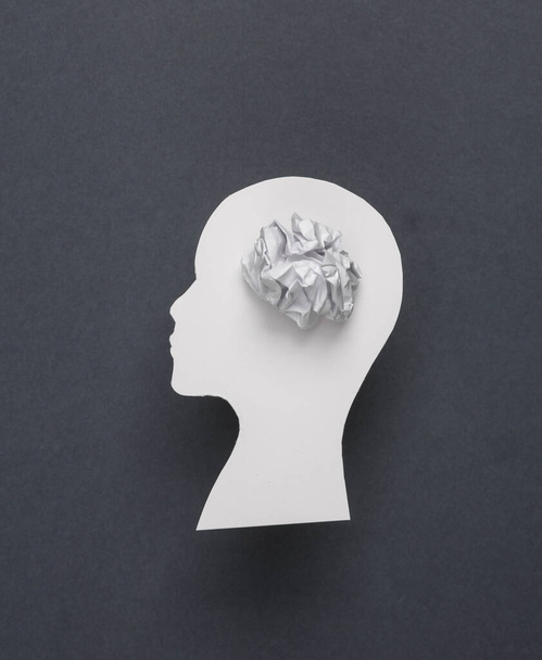 Mental health, brain disease, dementia, confused thoughts. Paper-cut silhouette of a human head with a crumpled piece of paper on a gray background - Photo, Image