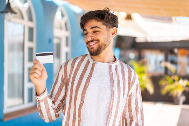 Yakışıklı bir Arap açık havada elinde mutlu bir ifadeyle kredi kartı tutuyor. - Fotoğraf, Görsel