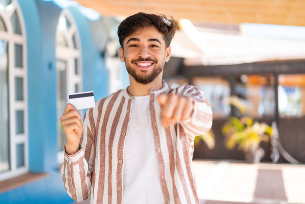 Açık havada kredi kartı tutan yakışıklı bir Arap, kendine güvenen bir ifadeyle seni işaret ediyor. - Fotoğraf, Görsel