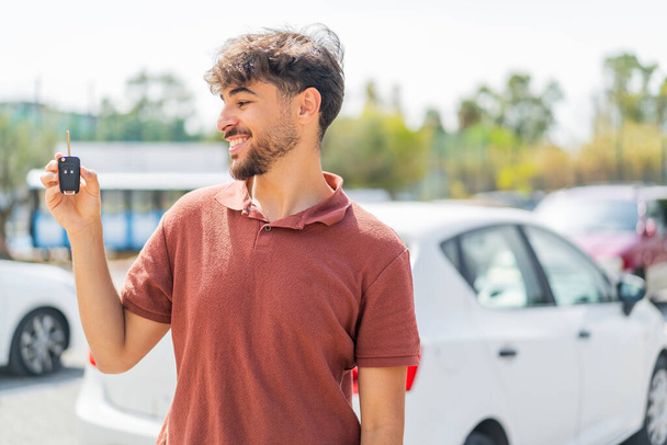 Νεαρός Άραβας όμορφος άνδρας κρατώντας το κλειδί του αυτοκινήτου σε εξωτερικούς χώρους με ευτυχισμένη έκφραση - Φωτογραφία, εικόνα