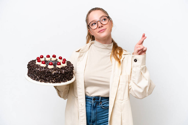 Joven mujer caucásica sosteniendo un pastel de cumpleaños aislado sobre fondo blanco con los dedos cruzando y deseando lo mejor - Foto, imagen