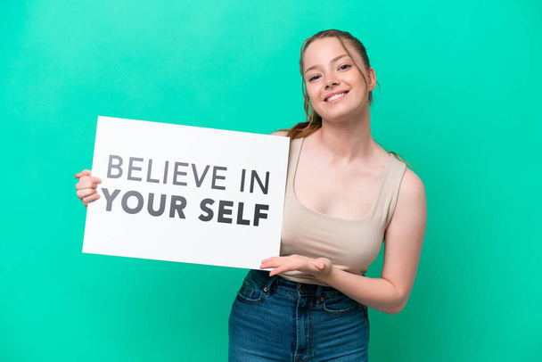 Молода жінка, ізольована на зеленому тлі, тримає плакат з текстом "Вірте у себе" і вказує на нього. - Фото, зображення