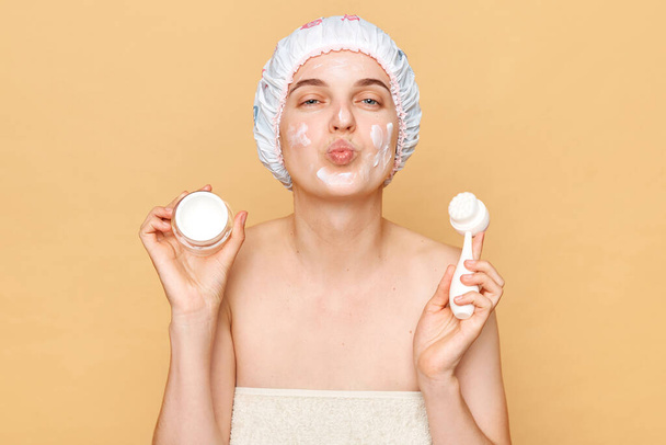 Beautiful woman wearing shower cap standing isolated over beige background making cleansing procedures for her face sending air kissing. - Zdjęcie, obraz