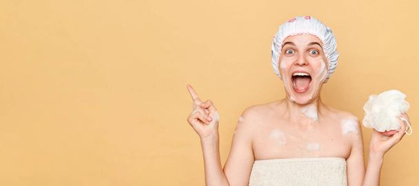 Amazed woman wearing shower cap standing isolated over beige background posing while taking shower pointing at copy space for promotion. - Photo, image