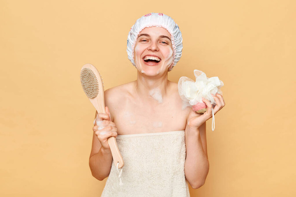 Positive laughing woman wearing shower cap standing isolated over beige background being in bath posing wrapped in towel holdingdry massage brush and washcloth. - Photo, image