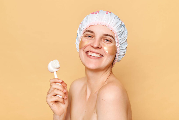 Cheerful woman wearing shower cap standing isolated over beige background enjoying skin care procedures after taking shower looking at camera with happy face. - Foto, Bild