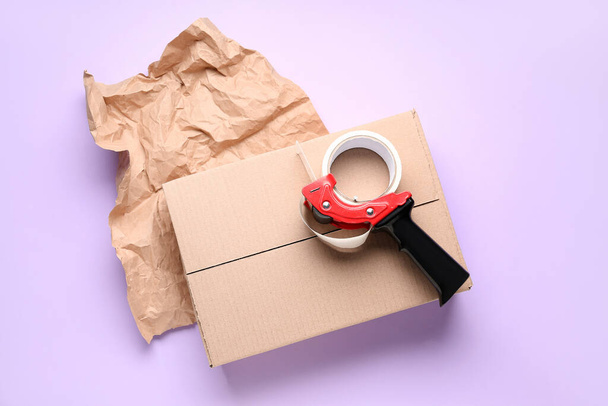 Dispensador de cinta de embalaje, caja de cartón y papel arrugado sobre fondo lila - Foto, Imagen