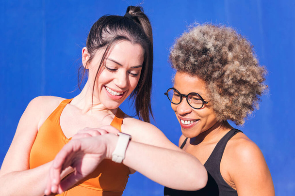 portrait of two multiethnic friends using a smart watch while practicing sport, concept of sports technology and active lifestyle, copy space for text - Foto, afbeelding