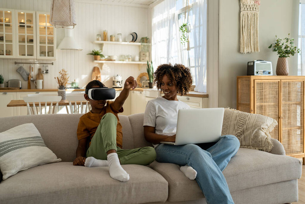 Афро-американская семья мама и сын сидят на диване с электронными устройствами. Мать-родитель сидит с ноутбуком, сын в очках виртуальной реальности. Чёрный парень показывает себя в игровом шлеме. Семья и технологии - Фото, изображение