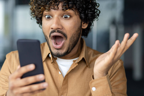 Close up of hispanic man received online win and win notification, man with curly hair holding smartphone. - Фото, изображение