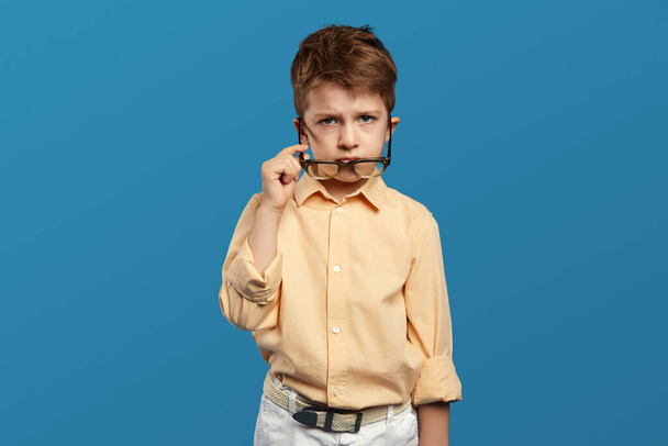 Zweifelhaftes kleines Kind im beigen Hemd, das durch eine Brille in die Kamera blickt. Verdächtiges Kind starrt isoliert auf blauem Hintergrund. - Foto, Bild