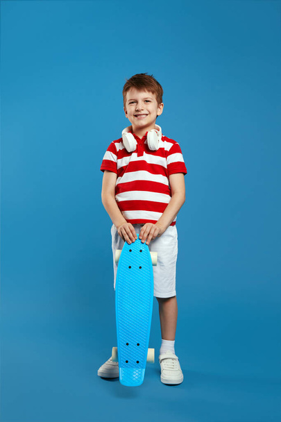 Vertical photo of full body excited kid boy wearing red striped polo shirt and headphones on neck, holding skateboard and smiling for camera, isolated over blue background - Photo, Image