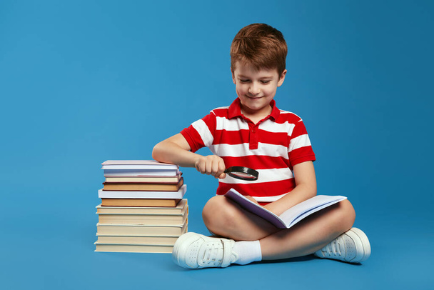 Gülümseyen ve kitap okumak için büyüteç kullanan sıradan giyinmiş mutlu bir çocuk bir sürü kitabın yanında, mavi arka planda izole bir şekilde oturuyordu. - Fotoğraf, Görsel