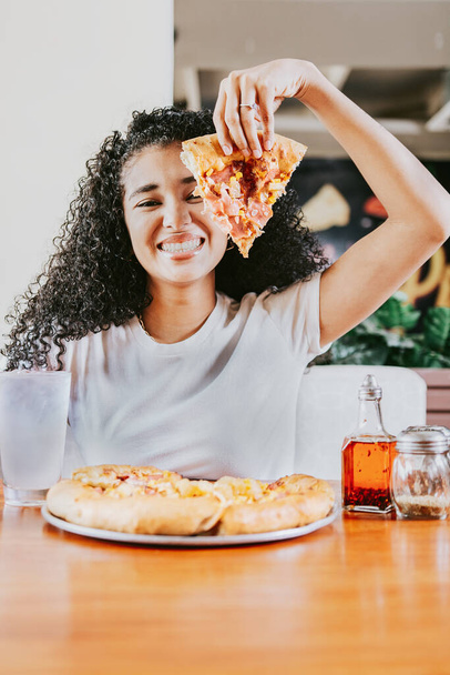 Bir restoranda pizza yerken gülümseyen afro kadın. Lokantada pizza gösteren mutlu afro saçlı kadın. - Fotoğraf, Görsel
