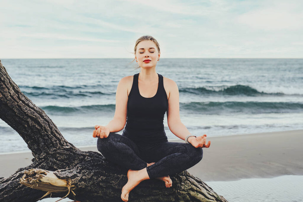 A girl in a black singlet and leggings meditates sitting on a snag on the shore of a lake or sea. - Photo, Image