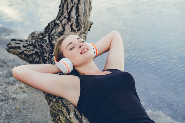 Dziewczyna w czarnej odzieży sportowej i dużej białej bezprzewodowej słucha muzyki leżąc na kłębku, uśmiechając się z zamkniętymi oczami. - Zdjęcie, obraz