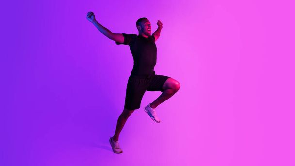 Sport victory, triumph. Emotional black runner man running raising hands and shaking fists in joyful gesture, celebrating win on purple neon background. Full length, free space for text. Panorama - Photo, Image