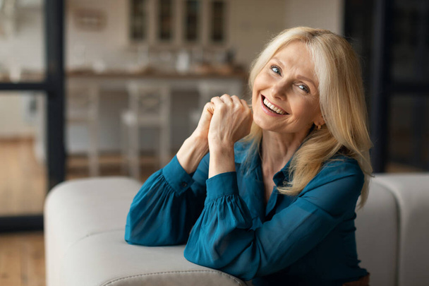 Portret van een vrolijke blanke volwassen vrouw die thuis op de bank rust, kijkend en glimlachend naar de camera. Vrolijke vrouw ontspannen in gezellige woonkamer, kopieer ruimte - Foto, afbeelding