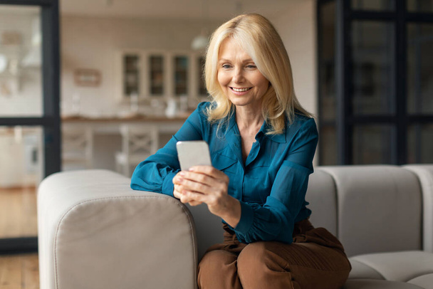 Smiling mature woman with smartphone sitting on couch at home, adult lady using mobile phone, messaging with family or browsing internet, purchasing in online web store - Foto, afbeelding