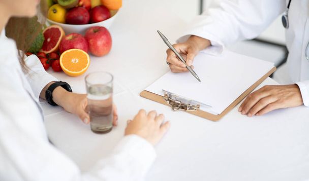 Europäische reife Ärztin Ernährungsberaterin im weißen Kittel schreibt Diätprogramm für die Behandlung von Patientin mit Wasser im Klinikinnenraum, Draufsicht. Gesundheitsfürsorge, Gewichtsabnahme mit professionellen - Foto, Bild