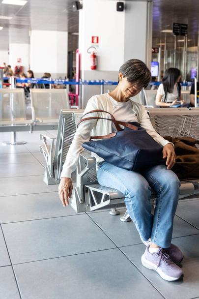 疲れたアジアの女性は,飛行を待っている間,空港出発ターミナル待機エリアで椅子で寝る - 写真・画像