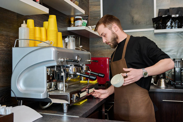 Male barista preparing coffee in coffee shop, bearded young man worker in an apron standing near coffee machine. Small business, food service occupation, coffee shop, staff, work concept - Fotografie, Obrázek