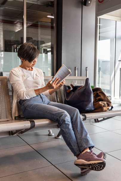 MIddle年齢 アジアの女性 空港ターミナルへの出発を待っている間に時間を殺すために荷物読書本 - 写真・画像