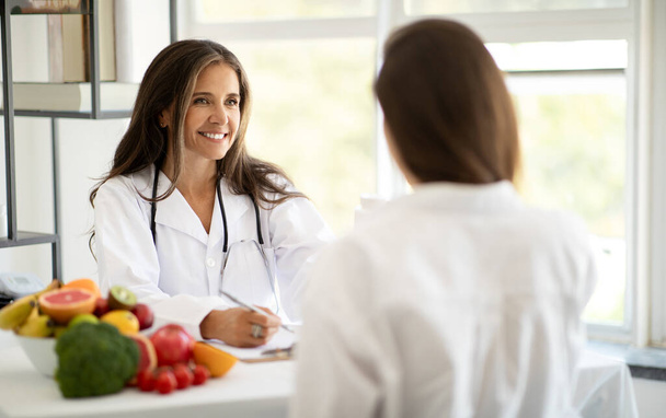 Cheerful european middle aged doctor nutritionist in white coat advises woman patient, recommends diet program in clinic interior. Treatment, weight loss with professional, health care - Photo, Image