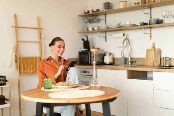 Cheerful young woman sitting in kitchen, using smartphone while having dinner, texting on phone, browsing social networks or shopping online, copy space - Fotó, kép