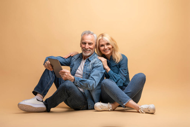 Positive beautiful loving senior spouses using digital tablet, isolated on beige background. Happy elderly man and woman paying for goods and services online, using banking or shopping app - Photo, Image