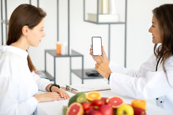 Heureuse femme européenne d'âge moyen médecin nutritionniste en manteau blanc conseille patiente, recommande app au téléphone avec écran vide à l'intérieur de la clinique. Gadget pour les soins de santé, traitement, minceur - Photo, image