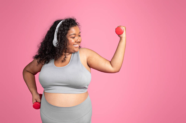 Молода афроамериканська жінка з позитивним тілом у спортивному одязі та бездротових навушниках займається вправами з ручними гантелями, насолоджується тренуванням з музикою на рожевому фоні студії, вільним простором - Фото, зображення