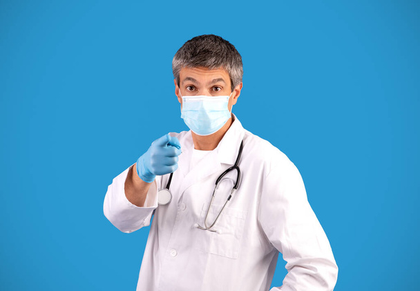 Γεια σου. Γκρι μαλλιά άνθρωπος γιατρός σε λευκό παλτό και μάσκα προσώπου δείχνοντας δάχτυλο στην κάμερα φορώντας γάντια από καουτσούκ στέκεται πάνω από το μπλε φόντο στούντιο. Ώρα για το ιατρικό σας ραντεβού - Φωτογραφία, εικόνα