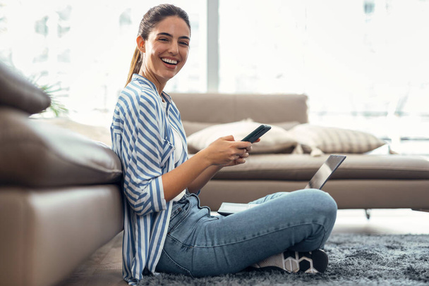 Съемка красивой молодой женщины с мобильного телефона во время работы с ноутбуком сидя на полу дома. - Фото, изображение