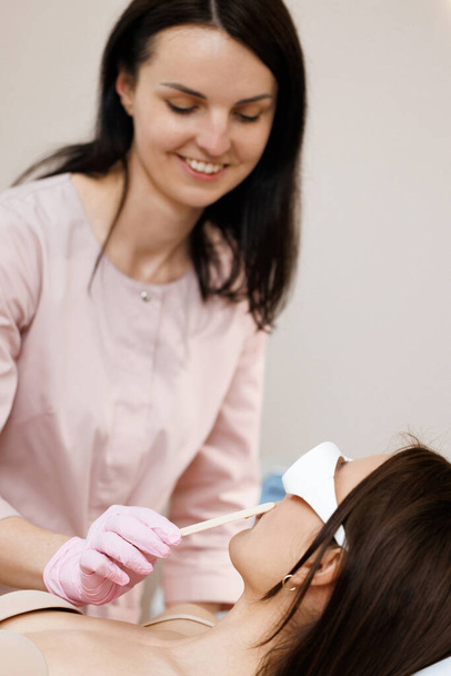 Beautician holds a wooden spatula and applies a transparent gel with anesthetic to the womans face and prepares the skin for the laser hair removal procedure. Laser mustache hair removal. - Photo, image
