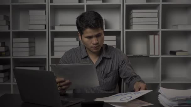 Asyalı erkek işadamı mali belgelerle ve masaüstü bilgisayarlarıyla geç saatlere kadar çalışıyor. Maaşlı çalışan genç adam konsepti 4K - Video, Çekim
