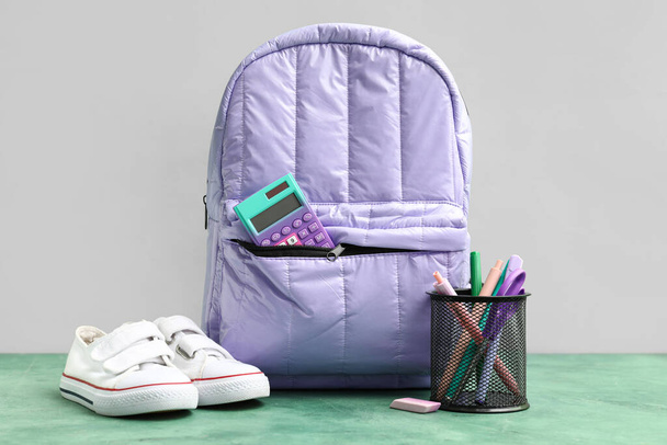 Рюкзак сирени с калькулятором, кроссовки и чашка ручек на зеленом столе возле белой стены - Фото, изображение