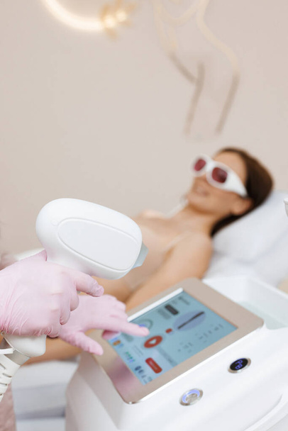Close up of female beautician in sterile gloves using diode laser hair removal machine while smiling woman laying on daybed. Esthetician preparing equipment for epilation procedure. - Фото, изображение