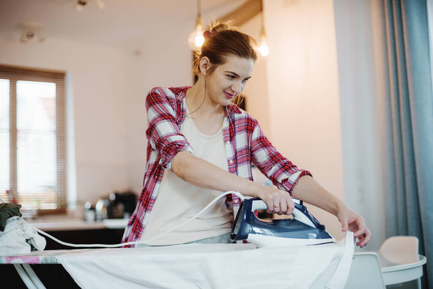 Νεαρή όμορφη γυναίκα σιδερώνει καθαρά ρούχα σε εσωτερικούς χώρους. Νεαρή γυναίκα σιδερώνει ρούχα. Χιονάτη μπλούζα. - Φωτογραφία, εικόνα