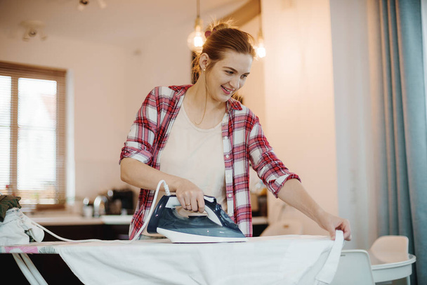 Νεαρή όμορφη γυναίκα σιδερώνει καθαρά ρούχα σε εσωτερικούς χώρους. Νεαρή γυναίκα σιδερώνει ρούχα. Χιονάτη μπλούζα. - Φωτογραφία, εικόνα