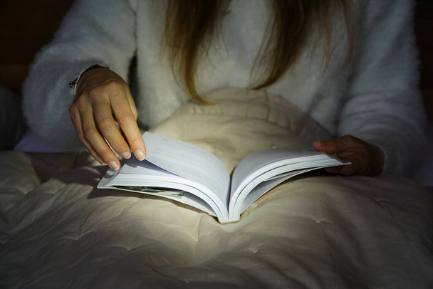 Image of a women reading a book in bed in the dark with a light on the center of the book. - Fotoğraf, Görsel
