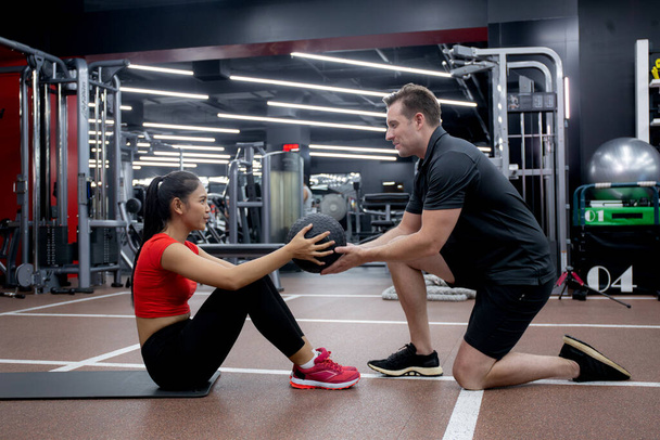 Молодая азиатская женщина и кавказский мужчина или тренер делает абс с мячом с решимостью вместе в фитнес-тренажерный зал, тренировки и упражнения на прочность и сильные мышцы, вес тренировки спортивная концепция. - Фото, изображение