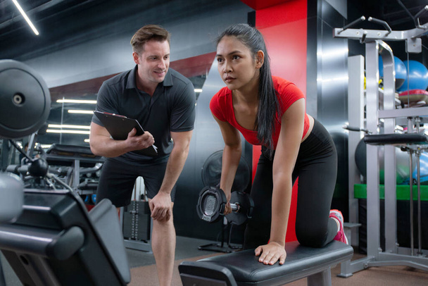 Kafkas erkek antrenör rehberliği ve Asyalı kadın spor salonunda motivasyon ve adanmışlıkla dambılı kaldırıyor, kaslı eller ve kollar için antrenman yapan kadın koç desteği, güç ve sağlık ile.. - Fotoğraf, Görsel