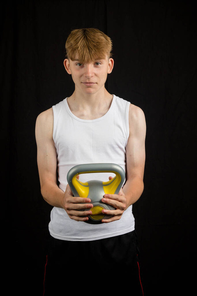 Portrait of a 14 year old caucasian teenage boy wearing a sleeveless vest holding a kettlebell against a black background boy - Fotó, kép
