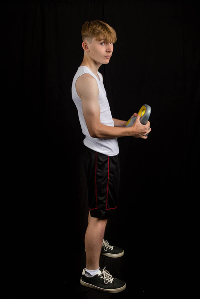 Portrait of a 14 year old caucasian teenage boy wearing a sleeveless vest holding a kettlebell against a black background boy - Фото, изображение