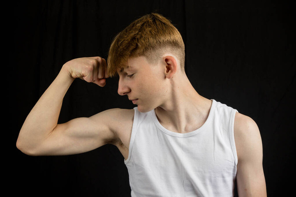 Portrait of a 14 year old caucasian teenage boy wearing a sleeveless vest flexing his bicep against a black background  - Foto, Imagem