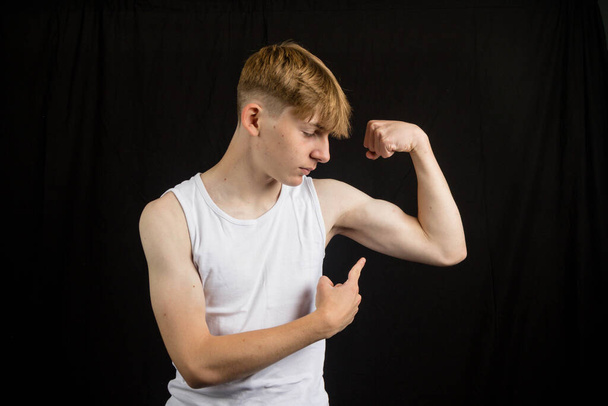 Portrait of a 14 year old caucasian teenage boy wearing a sleeveless vest flexing his bicep and pointing at it against a black background  - Photo, image
