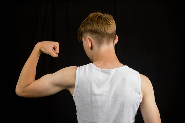 Portrait of a 14 year old caucasian teenage boy wearing a sleeveless vest flexing his bicep from behind against a black background boy - 写真・画像