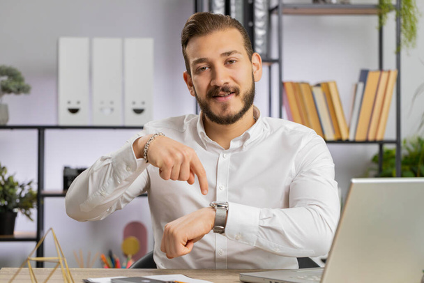 It is your time. Young middle eastern boss business man showing time on wrist watch clock, approve, pointing finger, time to start work, deadline. Freelancer guy at office workplace looking at camera - Photo, Image