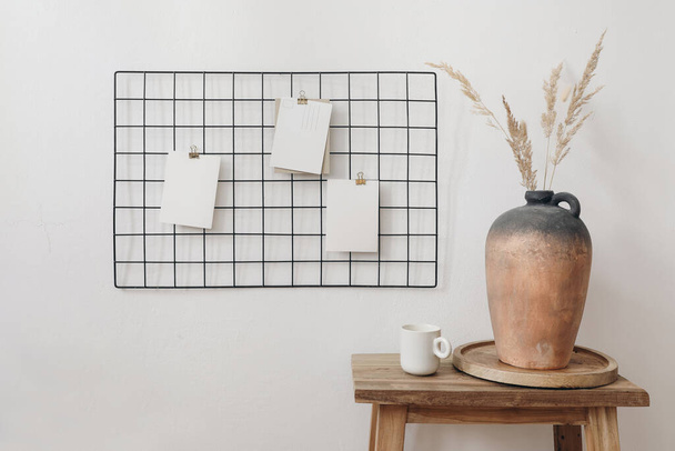 Black metal mesh noticeboard, bulletin board with blank memo cards mockups. Elegant home office interior concept. Clay vase with dry grass and cup of coffe on wooden table, white wall background. - Foto, Imagen